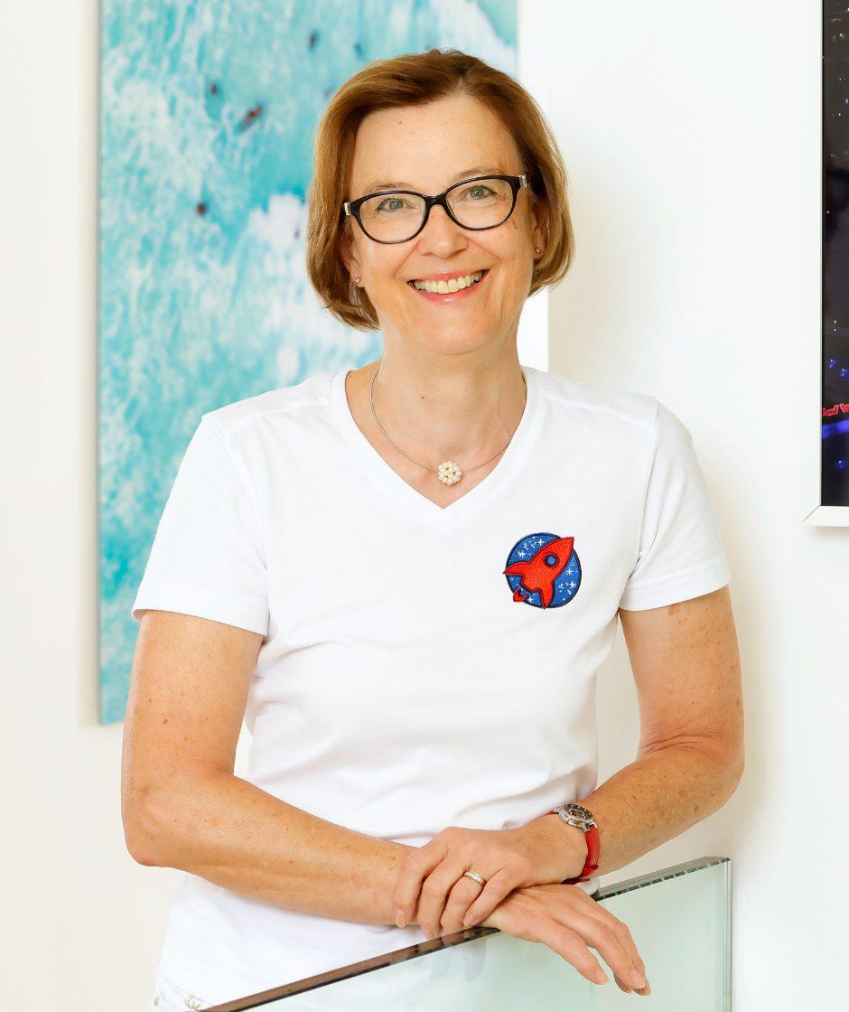 Portrait von Barbara Horvath in einer weißen Bluse. Sie lehnt auf einem gläsernen Geländer, im Hintergrund sieht man ein blaues Bild.
