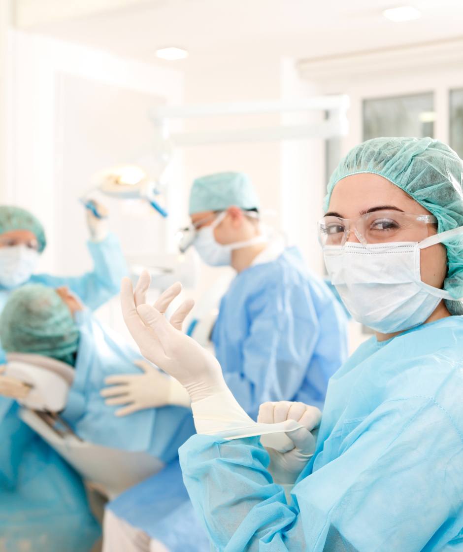 Dr. Horvath und zwei Assistentinnen führen gerade eine Operation im OP Zimmer durch. Alle tragen blaue OP Kittel und Schutzhauben auf dem Kopf.