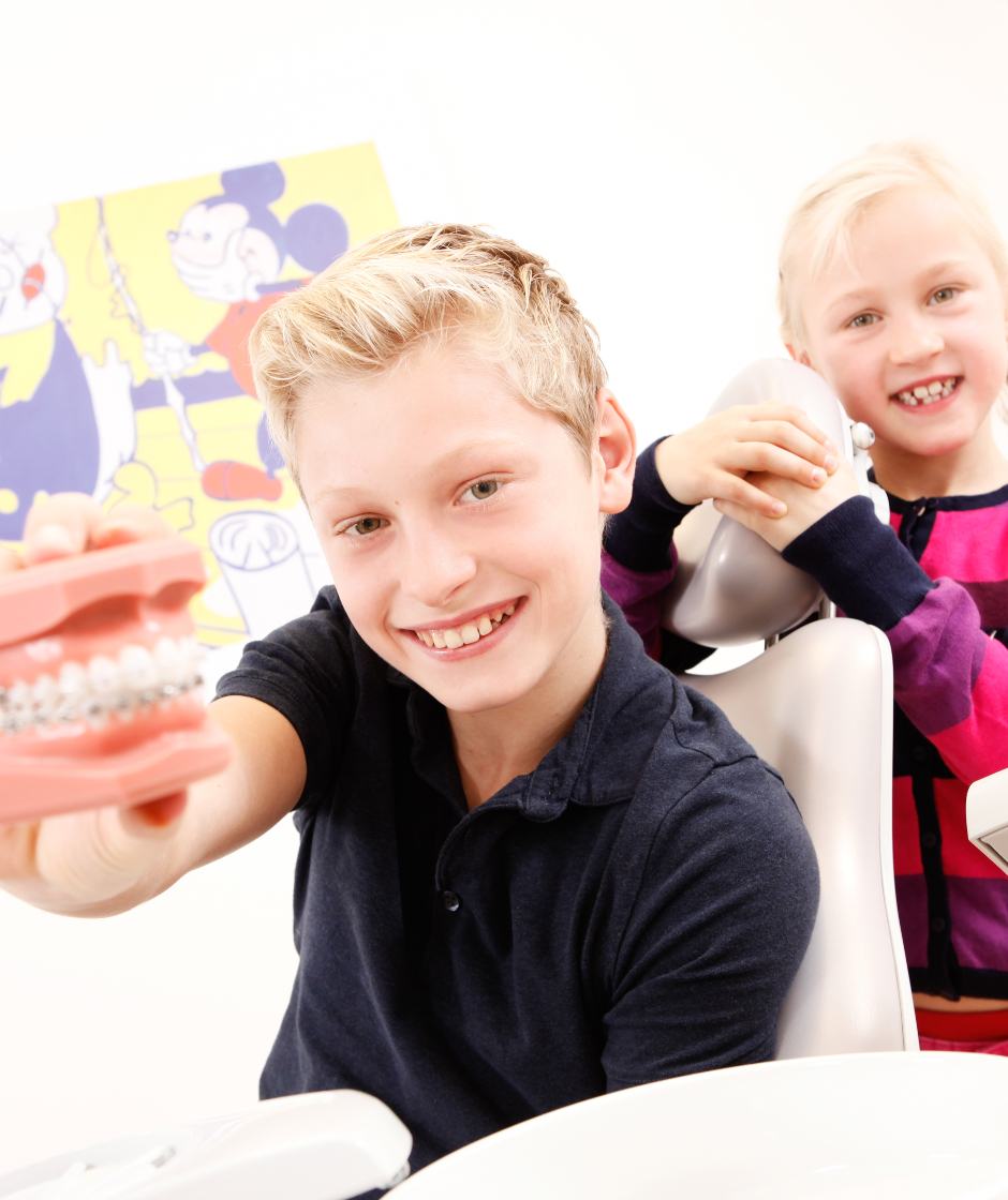 Zwei Kinder lächeln in die Kamera und halten ein Modell mit einer festen Zahnspange.