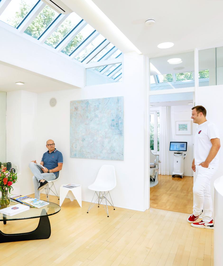 Lichtdurchfluteter Wartebereich mit Deckenfenstern. Dr. Sebastian Horvath begrüßt einen Patienten welcher gerade eine Zeitschrift ließt. Im Wartebereich steht ein Glaßtisch mit einem Blumenstrauß und Zeitschriften.