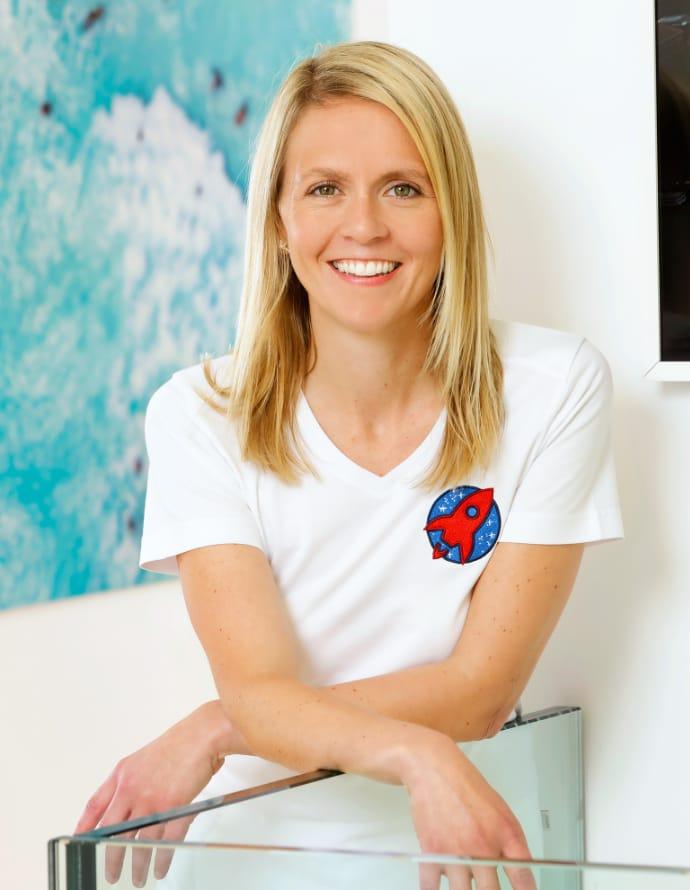 Portrait von Dr. Nicole Horvath, in einem weissen T-Shirt mit dem Praxislogo einer roten Rakete auf der linken Brust. Sie lehnt sich auf einem gläsernen Geländer, im Hintergrund sieht man ein blaues Bild.