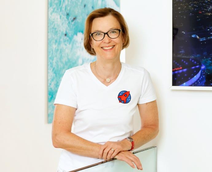 Portrait von Barbara Horvath in einer weißen Bluse. Sie lehnt auf einem gläsernen Geländer, im Hintergrund sieht man ein blaues Bild.