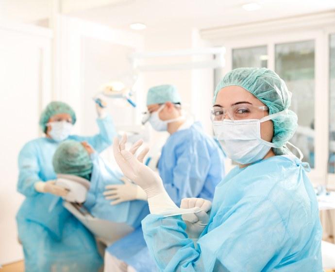 Dr. Horvath und zwei Assistentinnen führen gerade eine Operation im OP Zimmer durch. Alle tragen blaue OP Kittel und Schutzhauben auf dem Kopf.