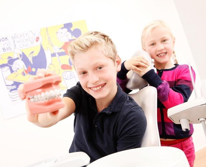 Zwei Kinder lächeln in die Kamera und halten ein Modell mit einer festen Zahnspange.