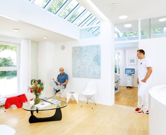 Lichtdurchfluteter Wartebereich mit Deckenfenstern. Dr. Sebastian Horvath begrüßt einen Patienten welcher gerade eine Zeitschrift ließt. Im Wartebereich steht ein Glaßtisch mit einem Blumenstrauß und Zeitschriften.