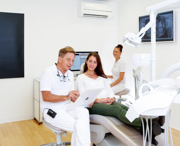 Dr. Domonkos Horvath bespricht einen Fall mit einer Patientin im Behandlungszimmer. Seine Assistentin ist im Hintergrund mit dabei.