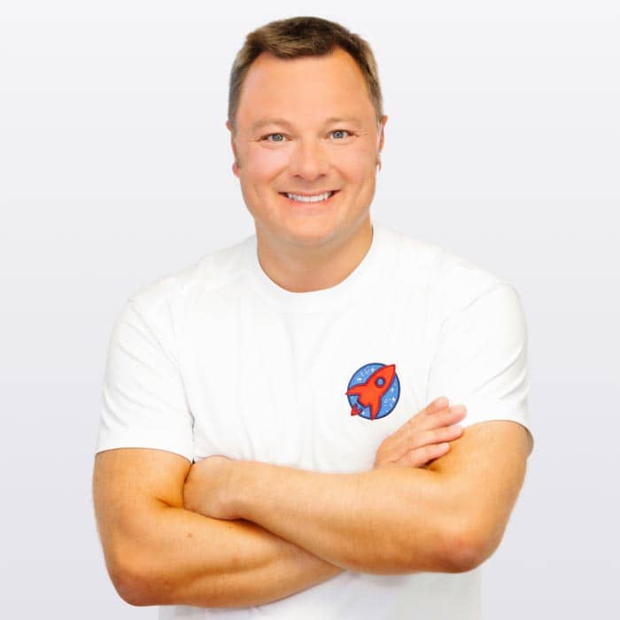 Professionelles Porträt von Dr. Sebastian Horvath, der mit verschränkten Armen lächelt. Seine zuversichtliche Ausstrahlung und das Praxislogo auf seinem T-Shirt repräsentieren das Engagement für exzellente zahnmedizinische Versorgung.