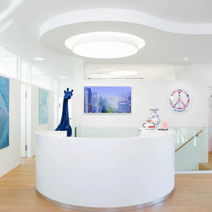 Helles und modernes Ambiente im Empfangsbereich von Dr. Horvaths Zahnarztpraxis: Die stilvolle Einrichtung mit kunstvollen Dekorationen und einer blauen Giraffenstatue schafft eine einladende Atmosphäre für Patienten.