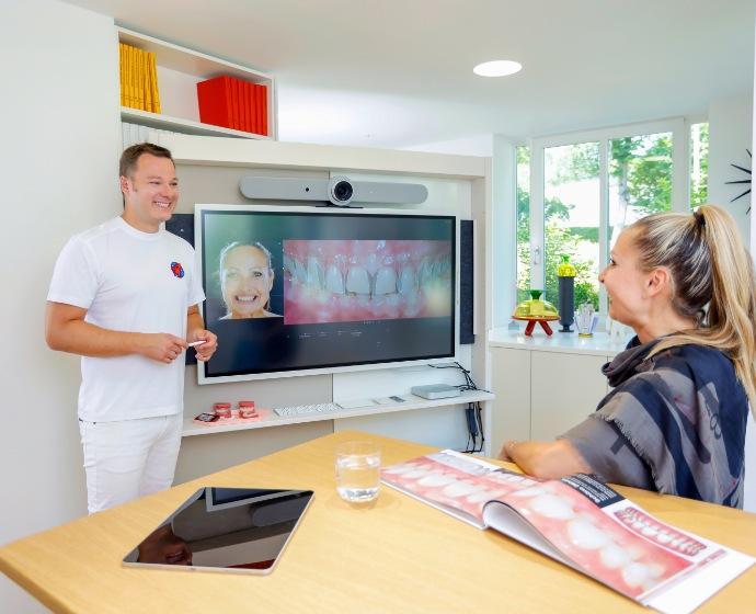 Dr. Sebastian Horvath präsentiert einer Patientin auf einem großen Bildschirm eine Simulation Ihres Behandlungsergebnisses mit Veneers. Die beiden befinden sich im Besprechungszimmer welches Lichtdruchflutet und mit verschiedener Kunst dekoriert ist.