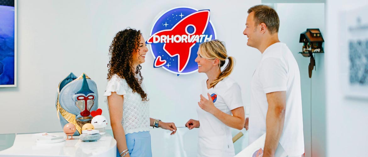 Dr. Nicole und Dr. Sebastian Horvath im Gespräch mit einer Patientin am Empfang, im Hintergrund das beleuchtete Raketen-Logo der Praxis.